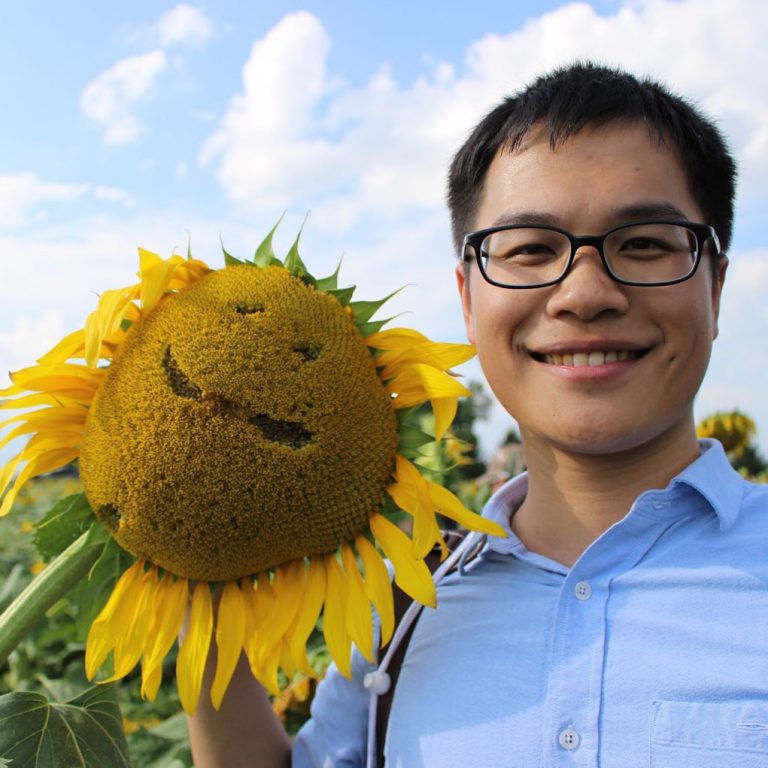 Photo of Min Fang with a sunflower to his left. The middle of the sunflower has a smiley face drawn into it.