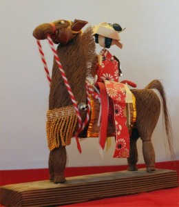 A woman, perhaps a bride, constructed of paper, rides a horse woven of some fine woody material, and decorated with silk cords and glass-headed pins. 